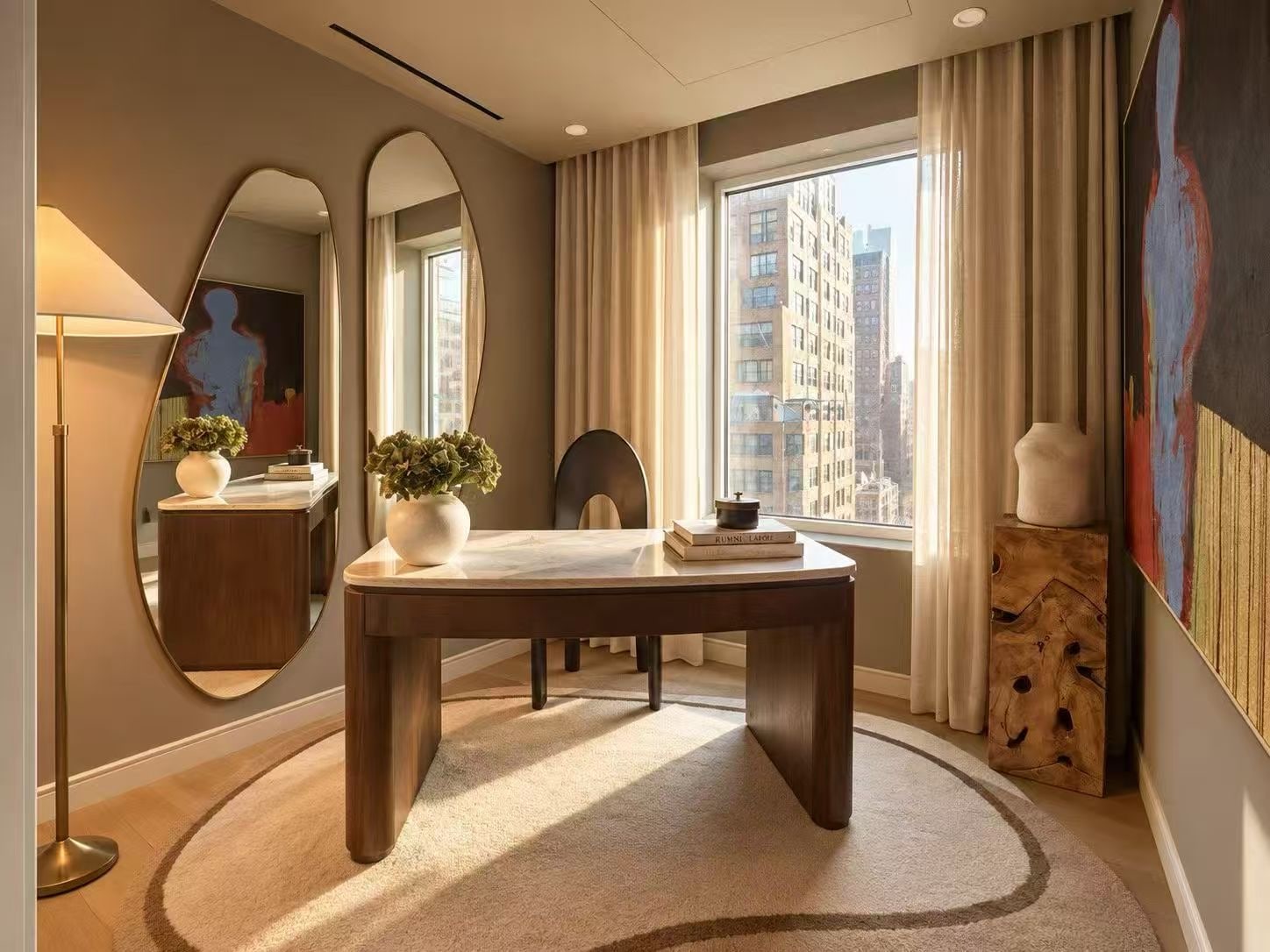 A home study with a walnut desk, oval mirror and a window onto neighboring towers